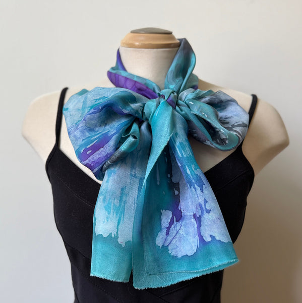 A lightweight silk scarf with an abstract watercolor pattern in shades of teal, blue, and purple, styled as a large bow around the neck of a mannequin wearing a black dress against a plain light gray background.